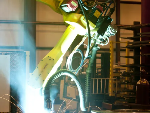 photo of a large, yellow machine welder arm, with a bright light at the welding site.