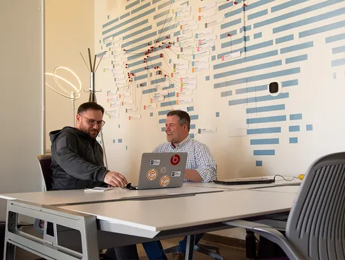 photo of two people seated at an office table in front of a wall with a stylized world map. One person has an open laptop, and the two people are engaged in a conversation.