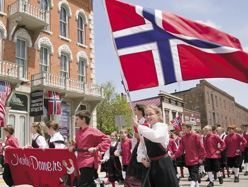 Children marching in a parade in traditional clothing and carrying a Norwegian flag