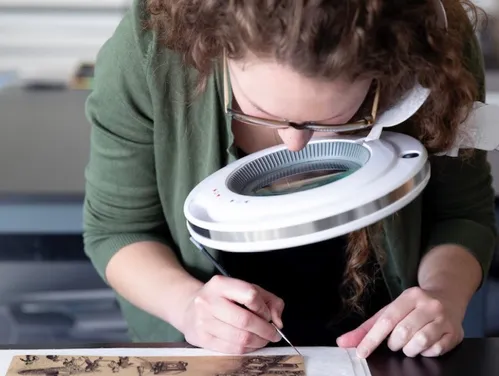 Erin Naylor works on restoring a paper object using a large magnifying glass