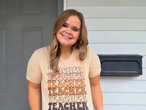 Female in teacher shirt standing in front of house.