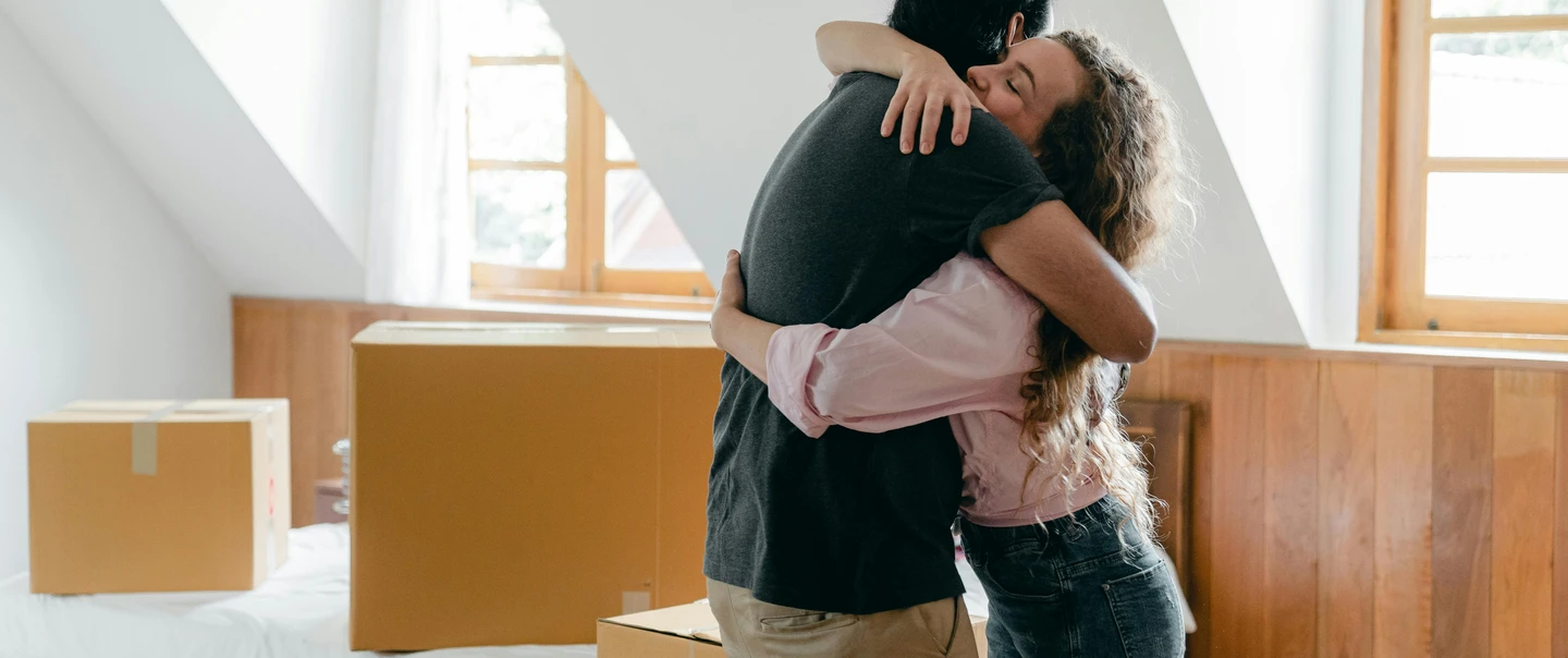 A man and woman hug in a white room, surrounded by moving boxes