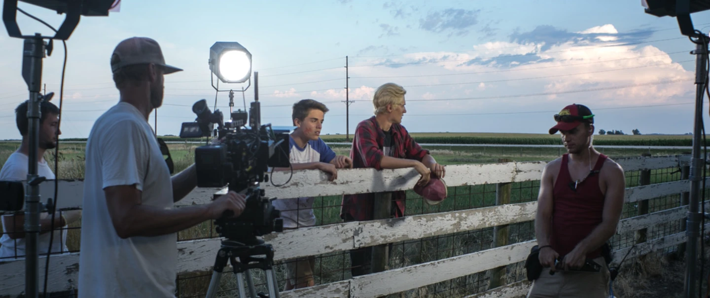 Five men film a movie in a field along a white fence