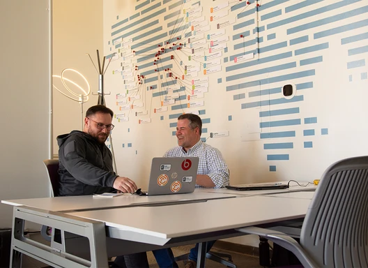 photo of two people seated at an office table in front of a wall with a stylized world map. One person has an open laptop, and the two people are engaged in a conversation.