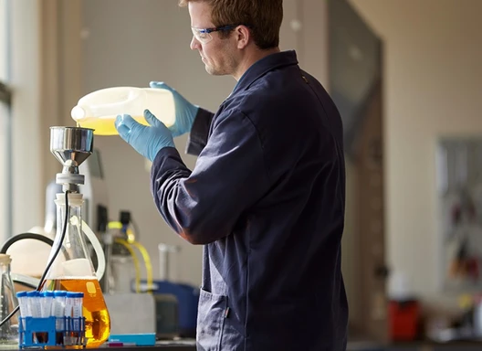 Man in coveralls pouring liquid into a large glass beaker