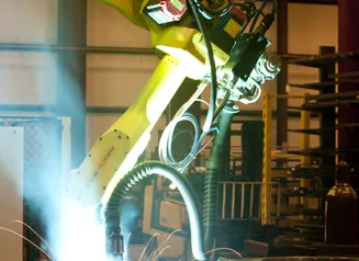 photo of a large, yellow machine welder arm, with a bright light at the welding site.