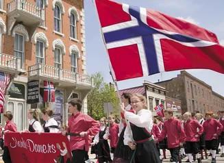 Children marching in a parade in traditional clothing and carrying a Norwegian flag