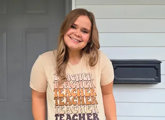 Female in teacher shirt standing in front of house.