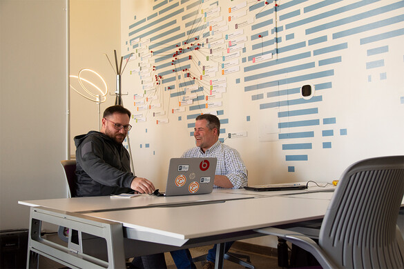 photo of two people seated at an office table in front of a wall with a stylized world map. One person has an open laptop, and the two people are engaged in a conversation.