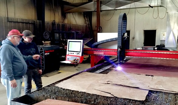 a photo of two people standing near a large CNC plasma cutter as it cuts a piece of material on a large bed beneath it.