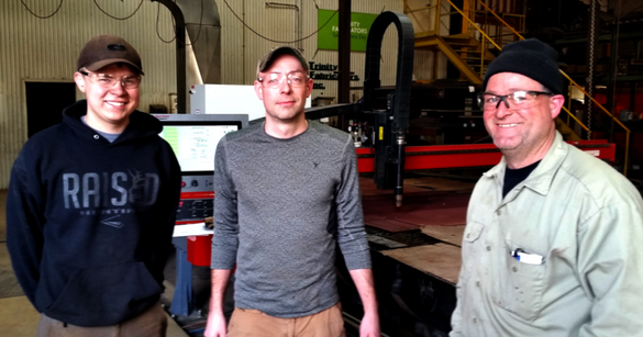 a photo of three people standing in front of the Trinity Fabricators logo in the manufacturing space. They are smiling and wearing some light protective gear.
