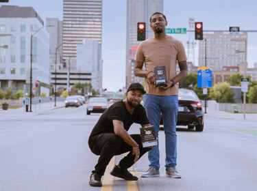 Pernell Cezar Jr. and Rod Johnson holding a bag of their coffee with downtown Des Moines behind them