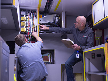 Two men working on on wires behind a panel
