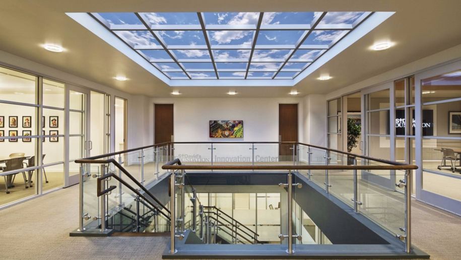 a photo of an office setting with a larger stairway in the middle. Above the stairs in an installation of ceiling panels that mimic a blue sky with white clouds.