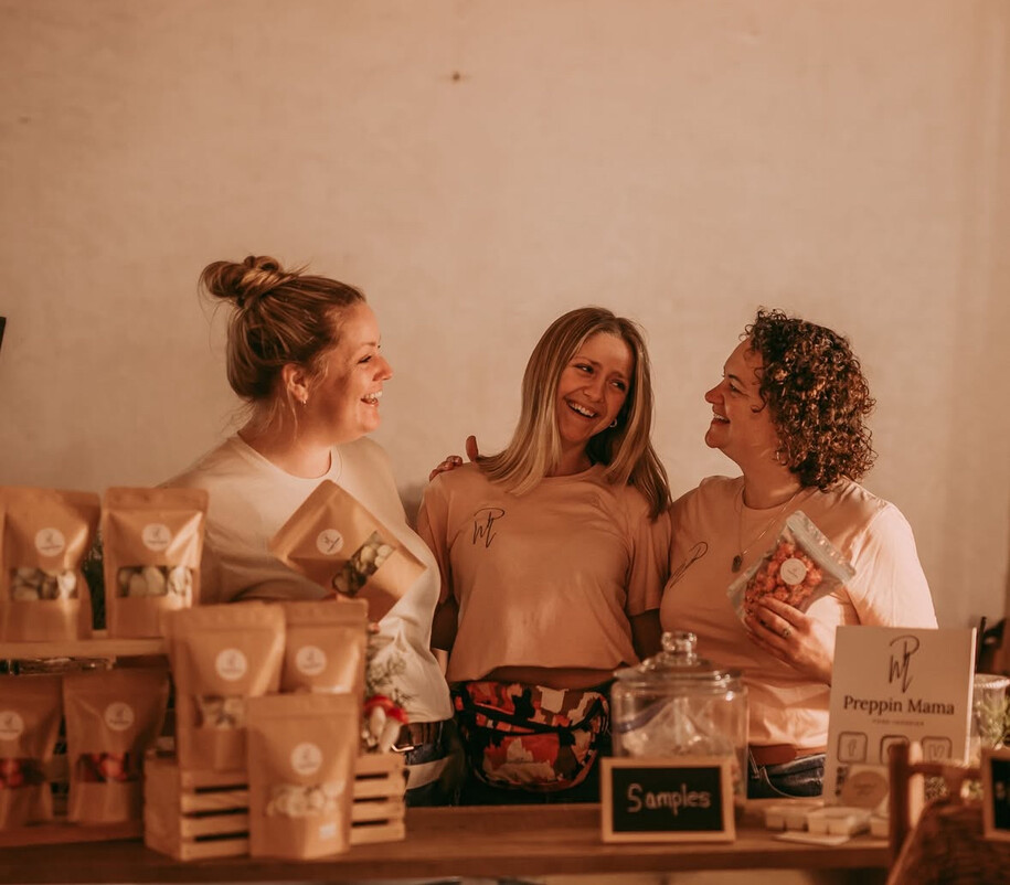 three women standing behind a counter with homemade snacks on it